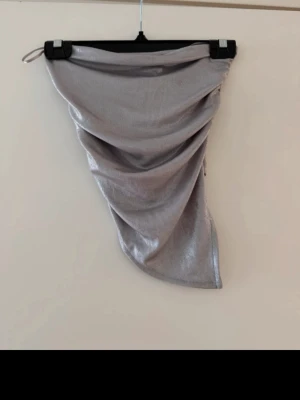 Silver glittrig bandeau tubtopp - Säljer en snygg silverfärgad bandeau tubtopp med glittrigt tyg. Toppen har en asymmetrisk form och är axelbandslös, perfekt för att sticka ut. Materialet är mjukt och följsamt med metallic finish.