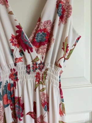 Vit långklänning med blommigt mönster - Säljer en vit långklänning med färgglatt blommigt mönster i rött, blått och grönt. Klänningen har v-ringning, breda axelband och smock i midjan för en bekväm passform. Perfekt för sommarens alla tillfällen!