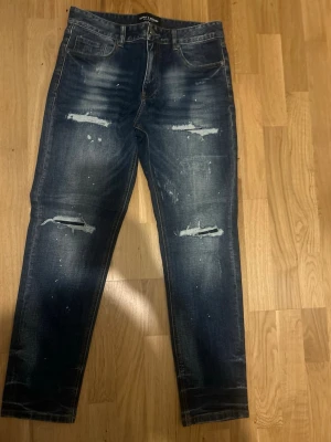 Mörkblå slitna supply&demand  jeans med hål - Säljer ett par mörkblå jeans med slitningar och hål på både lår och knän. Jeansen har en tvättad look med vita färgstänk och raka ben. Klassisk femficksmodell och knappgylf. Perfekta för en avslappnad streetstil.