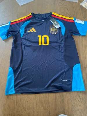Spaniens matchtröja Lamine Yamal #10 - Snygg mörkblå matchtröja från Adidas för Spaniens landslag med gula detaljer, nummer 10 och Lamine Yamal på ryggen. Tröjan har röda och gula ränder på axlarna, ljusblå ärmar och Climacool-material som andas. Perfekt för fotbollsträning eller att bära till matchen.