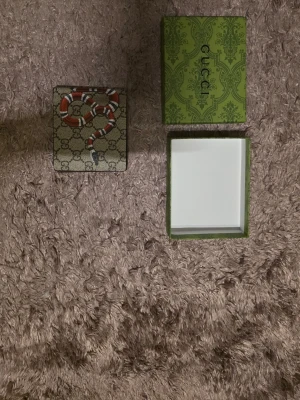 i plånbok med orm och-mönster - Säljer en snygg  plånbok i beige canvas med klassiskt -mönster och en röd, vit och svart orm på framsidan. Plånboken är fyrkantig och har en exklusiv känsla, perfekt för dig som gillar statement-accessoarer.