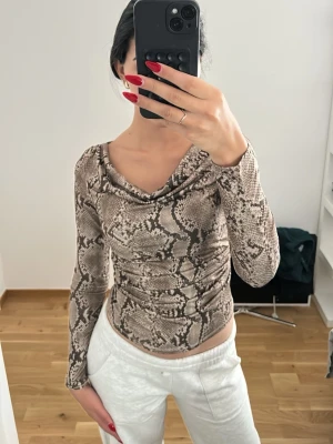 Brun långärmad topp med ormmönster - Säljer en brun och beige långärmad topp med trendigt ormmönster. Toppen har en snygg draperad urringning och är figurnära i modellen. Materialet är stretchigt och mjukt, perfekt för dig som vill ha en cool statement-piece i garderoben.