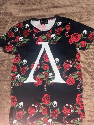 Svart t-shirt med rosor och dödskallar - Unik svart t-shirt från Slade Couture med stort vitt tryck framtill, täckt av röda rosor, dödskallar och ormar i rött och vitt. T-shirten har rund hals och korta ärmar. Perfekt för dig som gillar edgy och iögonfallande stil.
