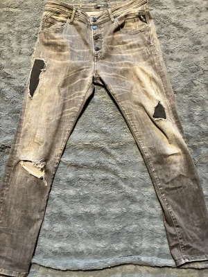 Replay - Säljer ett par grå jeans med slitningar och svarta patchdetaljer på benen. Jeansen har knappgylf, fem fickor och en smal passform. Snyggt tvättade med en vintagekänsla och råa kanter vid hålen. Perfekta för dig som gillar en edgy streetstil.