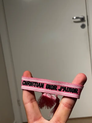 Christian Dior rosa tygarmband - Säljer ett rosa tygarmband från Christian Dior med texten 'Christian Dior J'Adior' i svart. Armbandet har tofsar i ändarna och är vävt i tyg med vita och rosa detaljer. Perfekt accessoar för dig som gillar lyxiga och trendiga detaljer.