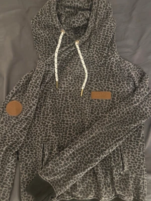 Grå leopardmönstrad hoodie - Snygg hoodie i grått med svart leopardmönster från fullSTAR. Har stor huva med vita snören och guldfärgade detaljer, samt två fickor framtill. Patch i brunt konstläder på bröstet och ärmen. Perfekt för dig som gillar djurmönstrat och streetstyle.