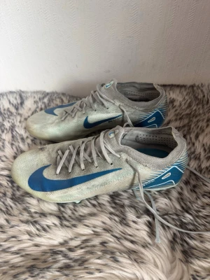 Nike ljusgrå-blå fotbollsskor - Säljer ett par Nike fotbollsskor i ljusgrått med blå detaljer och stor Swoosh-logga på sidan. Skorna har snörning, låg profil och dobbar i blått och vitt för bra grepp på planen. Tillverkade i syntetmaterial för lätt känsla och bra bollkontroll.