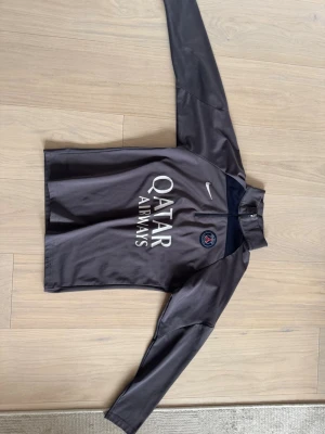Grå PSG träningströja från Nike - Snygg grå långärmad träningströja från PSG med halv dragkedja framtill. Tröjan har Paris Saint-Germain-logga på bröstet, Nike-logga och Qatar Airways-tryck. Tillverkad i ett lätt och funktionellt material som passar perfekt till fotbollsträning.
