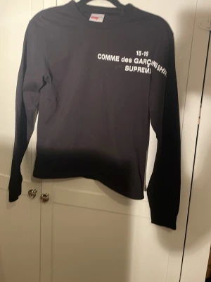 Svart långärmad topp Supreme x Cdg - Svart långärmad topp från Supreme x Comme des Garçons med vit text på bröstet. Klassisk crewneck och rak passform. Perfekt för dig som gillar streetwear och vill sticka ut med en unik collab.