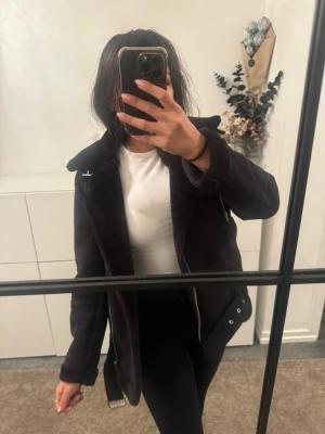 Svart jacka med bälte - Säljer en svart jacka med bälte och dragkedja. Jackan har lång ärm, bred krage och detaljer i silver. Perfekt för dig som vill ha en stilren och edgy look till hösten eller vintern.