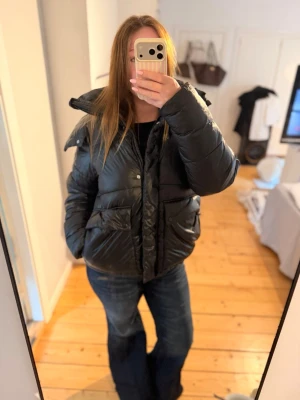 Oversize  pufferjacka - Säljer en svart oversize pufferjacka med stor huva och dragkedja framtill. Jackan har två stora fickor med lock och tryckknappar. Den är kortare i modellen och har en glansig finish som ger en cool vibe. Perfekt för kalla vinterdagar. Jackan är köpt här på plick. Storleken står inte men skulle tippa på en  storlek M.  Passar vem som beroende på hur oversize man vill ha den!