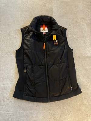 Svart dunväst från Parajumpers - Säljer en ÄKTA Snygg svart dunväst från Parajumpers med hög krage, dragkedja framtill och två fickor med dragkedja. Västen har en gul detalj vid halsen och klassisk Parajumpers-logga på bröstet. Perfekt för lager på lager och håller dig varm under kyliga dagar. Storlek S köpt på lantan outlet men passade ej aldrig använt den! 