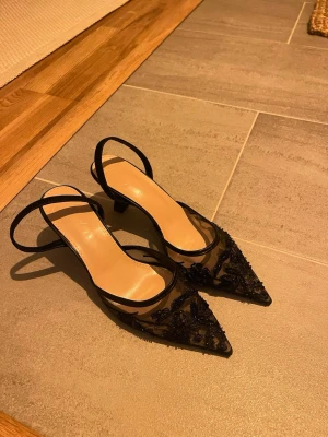 Svarta spetsiga pumps med mesh och pärlor - Säljer ett par eleganta svarta pumps med spetsig tå, låg klack och transparent mesh. Dekorerade med svarta pärlor och broderier för en lyxig känsla. Skorna har en öppen häl med smal rem runt ankeln. Perfekta för dig som vill sticka ut. Aldrig använda 