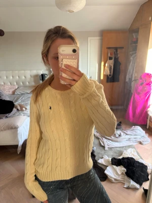 Gul kabelstickad tröja från Ralph Lauren - Kabelstickad gul tröja från Ralph Lauren med rund halsringning och lång ärm. Tröjan har den klassiska broderade loggan på bröstet och är tillverkad i ett mjukt stickat material. Perfekt för en avslappnad och stilren look. Den är i en ljusgul/beige färg 