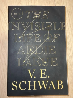 The Invisible Life of Addie LaRue - En roman med svart omslag och guldtext, skriven av V.E. Schwab. Bokens framsida har stjärnmönster och elegant typografi. Perfekt för dig som gillar fantasy och magisk realism. Språk: Engelska 