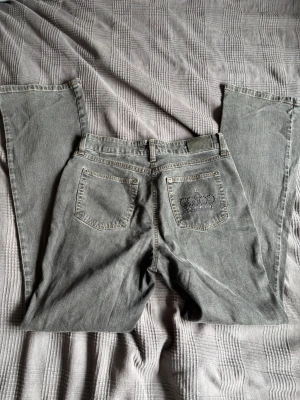 Grå jeans med krona på bakfickan - Säljer ett par grå jeans med raka ben och cool krona i nitar på ena bakfickan. Klassisk femficksmodell med bälteshällor och slitna detaljer för en avslappnad look. Perfekta för dig som vill sticka ut lite extra.
