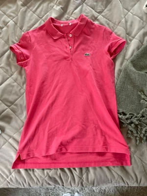 Korall pikétröja från Lacoste - Säljer en klassisk korall pikétröja från Lacoste med korta ärmar och krage. Tröjan har knappar framtill och den ikoniska krokodilloggan på bröstet. Perfekt för en sportig och stilren look.