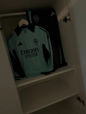 Ljusblå långärmad Arsenal träningströja - Snygg ljusblå långärmad träningströja från Arsenal med svarta detaljer och klubbmärke på bröstet. Tröjan har dragkedja vid halsen och Adidas-logga. Perfekt för dig som gillar fotboll och vill ha en sportig look.