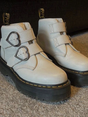 Vita Dr. Martens Devon heart platform skor, storlek 40 - Säljer ett par vita Dr. Martens plattåskor / platform boots med chunky svart sul och gula sömmar. Skorna har två breda remmar med coola hjärtformade spännen i silver. Tillverkade i kraftigt läder och har klassisk Dr. Martens-tag baktill. Originallåda.