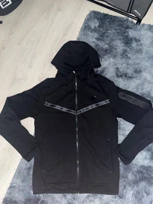 Hoodie och byxor Nike - Säljer en svart hoodie från Nike med hel dragkedja framtill och huva. Hoodien har diskreta grå detaljer över bröstet och på ena ärmen, samt fickor på sidorna. Perfekt för en sportig och avslappnad stil.