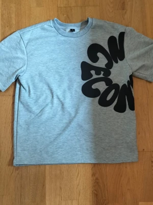 Grå t-shirt med stor svart text - Snygg grå t-shirt med stor, vågig svart text över bröstet och axeln. T-shirten har rund halsringning och korta ärmar. Perfekt för dig som gillar grafiska tryck och en avslappnad stil.