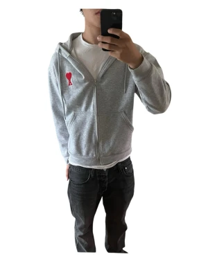 Ami Paris Zip - Modellen i bilden är 180cm och väger 69kg Snygg grå hoodie med dragkedja och huva, samt en röd hjärtformad patch på bröstet. Några små fläckar har den, men de märks inte. Hoodien har fickor framtill och justerbara snören i huvan. Perfekt för en avslappnad och trendig stil. Skriv vid minsta fundering. Skickar snabbt📦