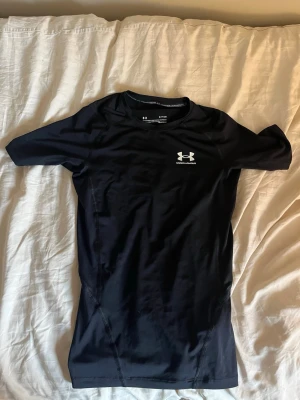 Svart träningströja Under Armour - Säljer en svart, tight och kortärmad träningströja från Under Armour. Tröjan har en vit logga på bröstet och är gjord i ett stretchigt funktionsmaterial som andas. Perfekt för gym, löpning eller annan sport.