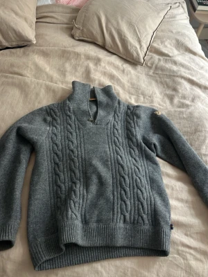 Fjällräven - kabelstickad half-zip - Mysig grå stickad tröja med kabelstickat mönster framtill och hög krage med halv dragkedja. Tröjan har långa ärmar och ribbade muddar vid ärmslut och nederkant. Perfekt för kalla dagar och enkel att styla till jeans.