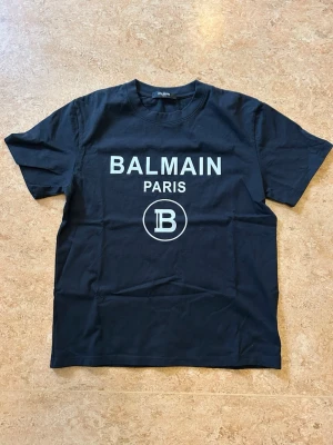 Svart Balmain Paris t-shirt - Snygg svart t-shirt från Balmain Paris med vit logga och text framtill. Klassisk rund halsringning och korta ärmar. Tillverkad i mjuk bomull med lite lycra för extra stretch. Perfekt för dig som vill ha en stilren och trendig look. Ny aldrig använd 