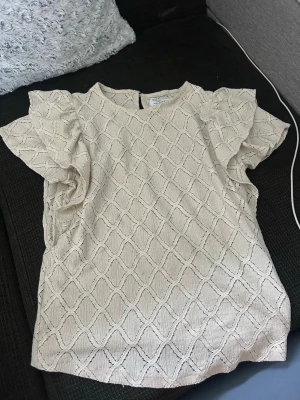 Beige blus med volangärmar - Snygg beige blus med strukturmönster och volangdetaljer vid ärmarna. Blusen har rund halsringning och ett diskret hålmönster i tyget som ger en trendig look. Perfekt att matcha med jeans eller kjol för en chic stil.
