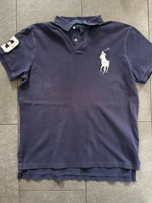 Mörkblå pikétröja från Polo Ralph Lauren - Klassisk mörkblå pikétröja från Polo Ralph Lauren med vit broderad logga på bröstet och siffran 3 på ärmen. Tröjan har krage, knappar framtill och är tillverkad i bomull. Perfekt för en sportig och stilren look. Gott skick men något urtvättad i färgen.