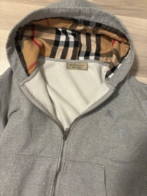 Grå zip hoodie från Burberry - Säljer en grå zip hoodie från Burberry med klassiskt rutigt foder i huvan.