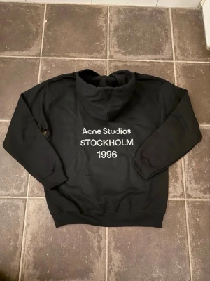 Svart hoodie Acne Studios Stockholm - Cool svart hoodie från Acne Studios med vit text 'STOCKHOLM 1996' på bröstet. Klassisk passform med huva och ribbade muddar. Perfekt för dig som gillar stilrena och trendiga plagg. Materialet är mjukt och bekvämt, perfekt för chill dagar. Acne är lite oversize men den passar som en vanlig M