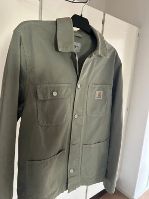 Ljusgrön overshirt från Carhartt - Snygg ljusgrön overshirt från Carhartt med fyra stora fickor framtill och klassisk krage. Tillverkad i slitstark bomull och stängs med knappar. Perfekt lager-på-lager-plagg för en avslappnad streetwear-look.