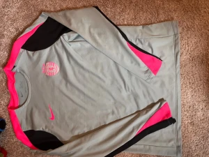 Nike långärmad träningströja PSG - Snygg långärmad träningströja från Nike i ljusgrått med svarta och neonrosa detaljer. PSG-logga på bröstet och Nike Swoosh i rosa. Tillverkad i Dri-FIT-material som andas och transporterar bort fukt. Perfekt för fotbollsträning eller gym.