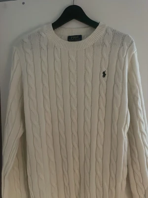  Polo Ralph Lauren - Kabelstickad vit tröja från Polo Ralph Lauren med rund halsringning och klassisk broderad logga på bröstet. Tröjan har långa ärmar och ribbade muddar vid ärmslut och nederkant. Perfekt för en clean och stilren look.