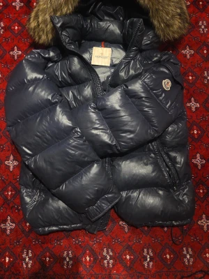 Moncler Maya Fur (ÄKTA – Limited edition  - Säljer min mörkblå Moncler Maya Fur (Mayaf) dunjacka då den inte används längre. Detta är lyxmodellen med tillhörande pälskrage på luvan(avtagbart).  Köpt för 17 999 kr. Jackan är använd men i fint skick. QR-kod/Certilogo på insidan som tar dig direkt till Monclers officiella hemsida. Storlek 4/ L.                                                        Jag har ägt den i två år, men under åtta av de månaderna låg jag inne i militären. Jackan har därav använts sparsamt 