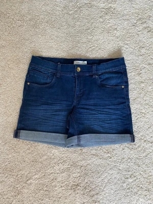 Mörkblå denimshorts - Snygga mörkblå jeansshorts med klassisk femficksdesign och uppvikta benslut. Shortsen har normal passform och midja, samt bälteshällor och knappstängning. 