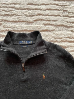 Mörkgrå halvzip från Polo Ralph Lauren - Mörkgrå långärmad tröja från Polo Ralph Lauren med halvzip och klassisk broderad logga på bröstet. Tröjan har en mjuk och skön känsla, ribbade muddar och hög krage. Perfekt för lager på lager och stilrena outfits. Inga skavanker. Längd: 67cm. Bredd: 56cm
