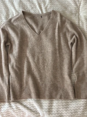 Beige stickad tröja med v-ringning  - Mysig beige stickad tröja med v-ringad hals och långa ärmar. Tröjan har bred ribbstickad kant nedtill och vid ärmsluten. Perfekt för lager på lager och enkel att matcha till olika outfits.