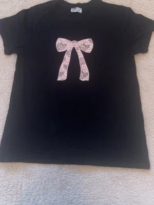 Svart t-shirt med rosa rosett - Svart t-shirt från Tyskland FB Sister med regular fit och ett stort tryck av en rosa spetsrosett på bröstet. T-shirten har korta ärmar och rund halsringning. Perfekt för dig som gillar söta detaljer och enkel stil.