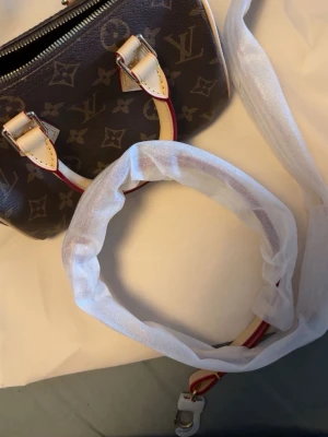 Louis Vuitton Monogram handväska - Säljer en klassisk Louis Vuitton handväska i brunt monogrammönster med detaljer i beige och rött läder. Väskan har två handtag och avtagbar axelrem. Kommer med dustbag och originalförpackning. Perfekt för dig som gillar lyxiga accessoarer.