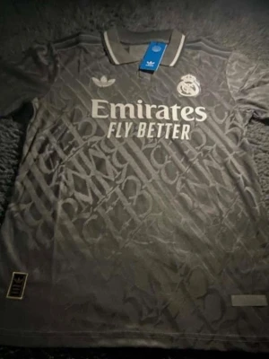 Grå jersey  - Snygg grå matchtröja från Real Madrid med  klubbmärke på bröstet. Tröjan har ett diskret mönster, klassisk krage och trycket 'Emirates Fly Better' på framsidan. Perfekt för fotbollsträning eller att bära till vardags.