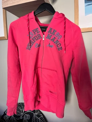 Rosa zip-hoodie från Peak Performance - Snygg rosa hoodie från Peak Performance med dragkedja framtill, huva med dragsko och känguruficka. Tryck i blått och vitt på bröstet samt liten logga vid fickan. Perfekt för en avslappnad och sportig stil.