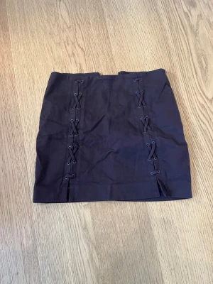 Mörkbrun kortkjol med snörning - Snygg brun kortkjol med cool snörningsdetalj framtill och dragkedja bak. Kjolen har en rak passform och är perfekt för dig som vill ha en edgy look. Tillverkad i ett mjukt material som känns bekvämt mot huden.