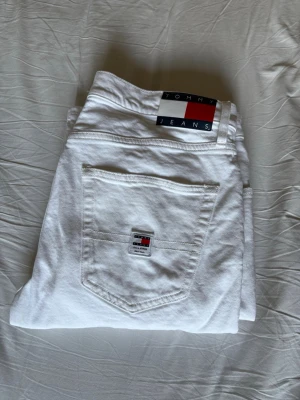 Vita jeans från Tommy Jeans - Säljer ett par fräscha vita jeans från Tommy Jeans med klassisk femficksmodell och tydlig logga på bakfickan samt linningen. Snygg passform och perfekt till dig som vill sticka ut med stilrena jeans.