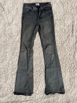 Grå bootcut jeans - Ett par grå bootcut jeans. Jeansen har slitningar och råa kanter nertill för en cool, avslappnad look. Materialet är denim i bomull med lite stretch för extra komfort.