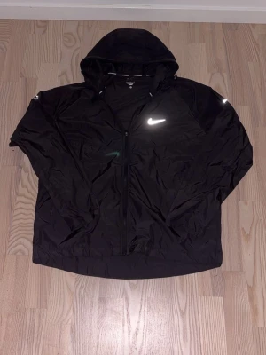 Svart vindjacka från Nike Running - Snygg svart vindjacka från Nike Running med klassisk vit Swoosh-logga på bröstet. Jackan är tillverkad i lätt polyester och har en sportig look med diskreta sömmar. Perfekt för löprundan eller när du vill ha en stilren och funktionell jacka.