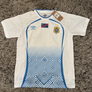 DR Kongo fotbollströja från Umbro - Säljer en vit och blå fotbollströja från Umbro med DR Kongos flagga och FECOFA-emblem på bröstet. Tröjan har korta ärmar, blå geometriskt mönster nedtill och blå detaljer längs sidorna. Perfekt för dig som vill stötta landslaget på match eller träning.