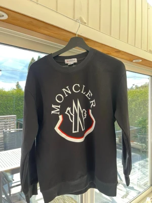Svart sweatshirt från Moncler - Snygg svart sweatshirt från Moncler med stort vitt och rött logotryck på bröstet. Tröjan har rund halsringning, ribbade muddar vid ärmslut och nederkant samt en Moncler-patch på ärmen. Perfekt för dig som gillar streetwear och vill ha något stilrent.